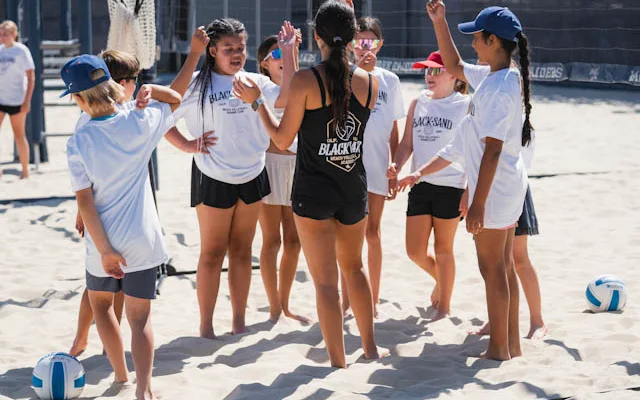 Campers in white Black Sand camp shirts huddle on the sand at summer camp