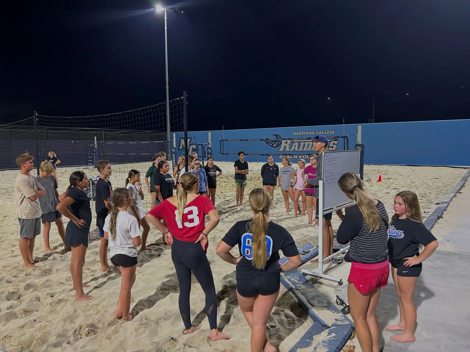 Young players gather around a coach at a whiteboard on the floodlit Moorpark College beach courts at night